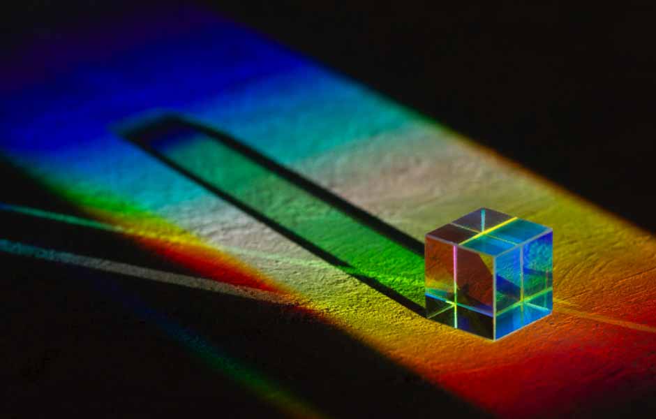Görselde, güneşten gelen beyaz ışığı mavi, kırmızı, sarı ve turuncu renklere dönüştürmüş parlak bir prizma var.