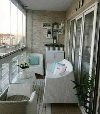 Cam Balkon Dekorasyonu Fikirleri. Görselde, cam sehpa, saksı çiçek, beyaz koltuklar ve duvar rafları olan minimalist tarzda bir balkon var.