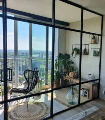 Cam Balkon Dekorasyonu Fikirleri. Görselde, içinde örgü sandalye, yer saksıları ve duvarında raf bölmeleri olan metal çerçeveli bir cam balkon var.