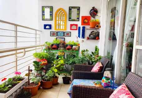 Görselde, beyaz duvarlarında özel dekorasyon ürünleri asılı ve zemininde çiçek saksılar olan ferah bir balkon var.