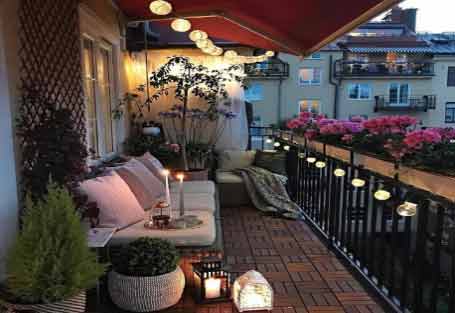 Geniş balkon dekorasyon fikirleri için kullanılan görselde, ahşap zemini, aydınlatma sistemi ve koltuk takımı ile modern bir geniş bir balkon var.