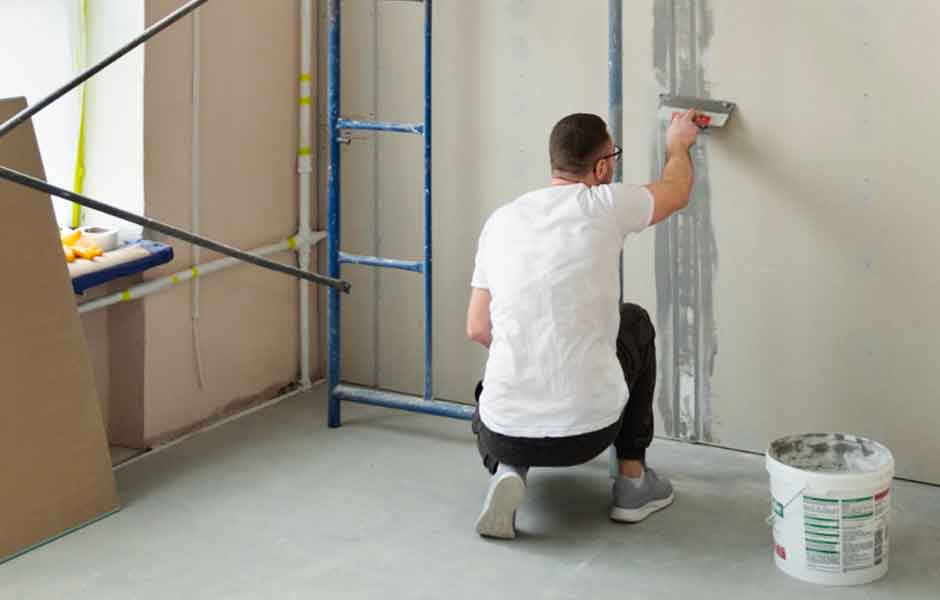 Görselde, duvar tamirinde uzmanlaşmış, duvara alçı süren profesyonel bir boyacı ustası var.