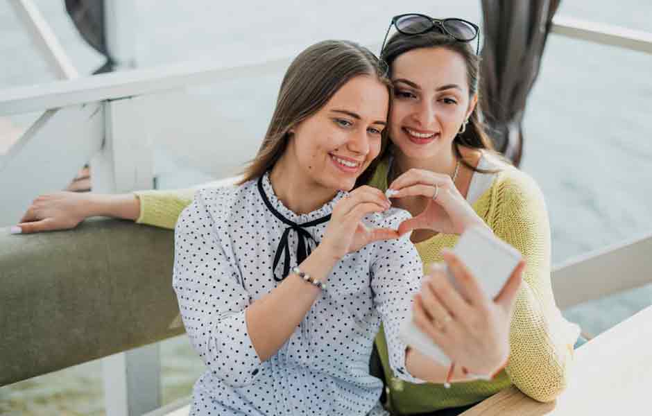 Anneler Günü için Güzel Sözler yazımız için kullandığımız görselde, iki genç kadın yeni anne olmuş arkadaşına telefonda görüntülü konuşuyor ve kalp işareti yapıyor.