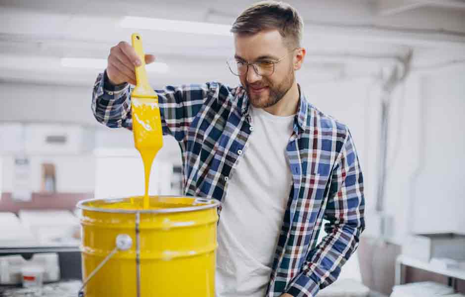 Görselde, boyacılık eğitimi alan genç adam, fırçayı içi sarı renkli boya ile dolu bir kovaya daldırıyor.
