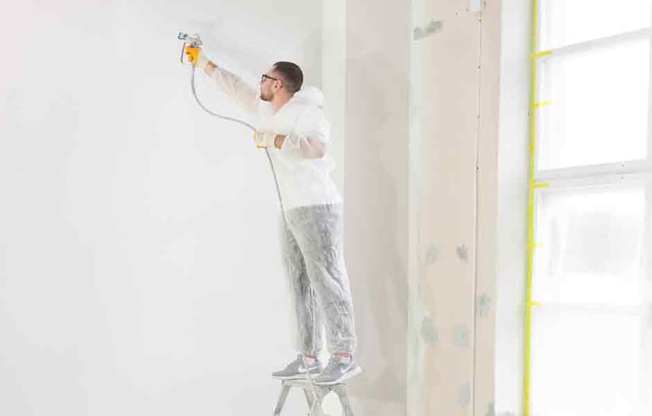 Görselde, profesyonel boyacı sprey tabancası ile salon duvarına boya püskürtüyor.